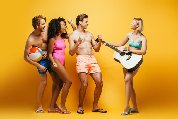 vista completa de la mujer rubia tocando la guitarra cerca de amigos multiculturales emocionados en traje de verano en amarillo - Foto, imagen