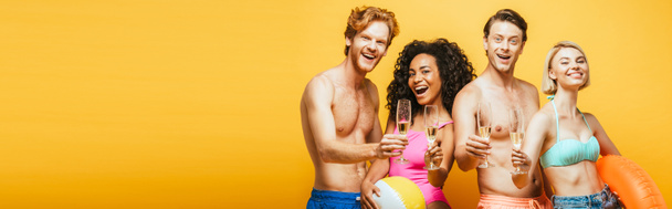 horizontální snímek mladých multikulturních přátel držících sklenice šampaňského, nafukovací kroužek a míč při pohledu na fotoaparát izolovaný na žluté - Fotografie, Obrázek