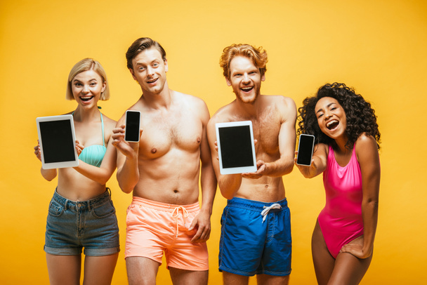 young multiethnic friends in summer outfit showing digital devices with blank screen while looking at camera isolated on yellow - Zdjęcie, obraz