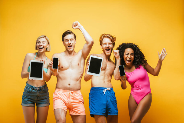 ενθουσιασμένοι πολυεθνικοί φίλοι με καλοκαιρινή ενδυμασία που δείχνουν ψηφιακές συσκευές με λευκή οθόνη στο κίτρινο - Φωτογραφία, εικόνα