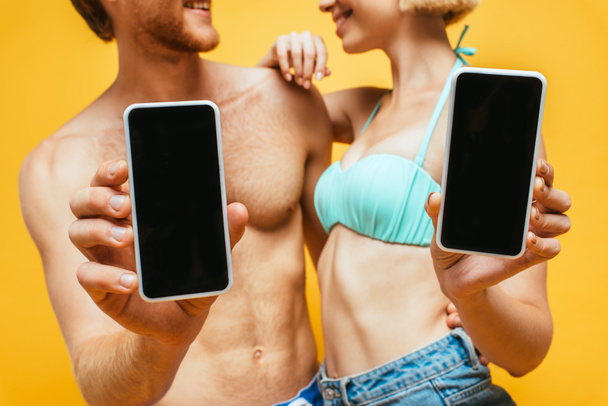 przycięty widok bez koszulki mężczyzna i kobieta w biustonosz pływacki pokazujący smartfony z pustym ekranem izolowane na żółtym, selektywnym ostrości - Zdjęcie, obraz