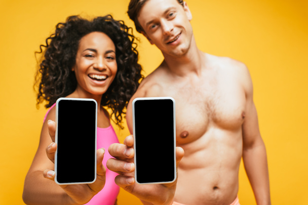 επιλεκτική εστίαση του γυμνού άνδρα και της αφρικανικής αμερικανικής γυναίκας που δείχνει smartphones με λευκή οθόνη απομονώνονται σε κίτρινο - Φωτογραφία, εικόνα