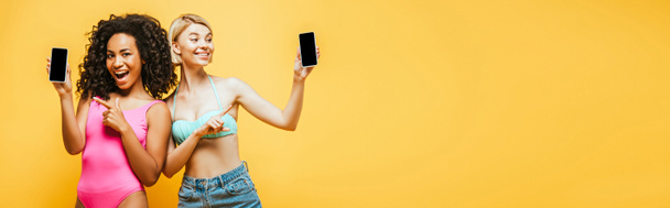 website header of young interracial women in summer outfit pointing at smartphones with blank screen isolated on yellow - 写真・画像
