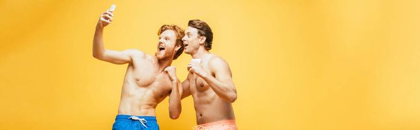 horizontales Bild von zwei Freunden ohne Hemd, die grimmig sind und geballte Fäuste zeigen, während sie ein Selfie mit dem Smartphone machen, isoliert auf gelb - Foto, Bild