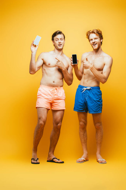 Ganzkörperansicht von zwei Freunden ohne Hemd in kurzen Hosen, die den Daumen nach oben zeigen und mit dem Finger zeigen, während sie Smartphones auf gelb halten - Foto, Bild
