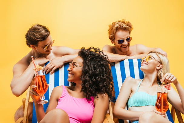multicultural women sitting in deck chairs with cocktails near shirtless men in sunglasses isolated on yellow - Photo, Image