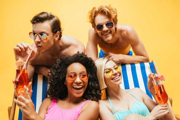 young shirtless men in sunglasses having fun with multicultural women sitting in deck chairs with cocktails isolated on yellow - Photo, image
