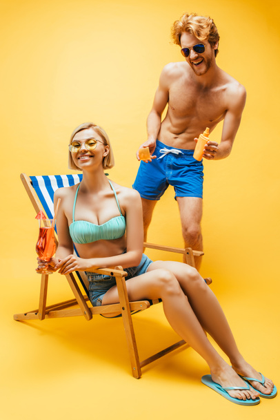 blonde woman sitting in deck chair with cocktail glass near man standing with sunscreen on yellow - Foto, imagen