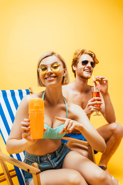 blonde woman sitting in deck chair and pointing with finger at orange juice near young man with cocktail glass isolated on yellow - Photo, Image