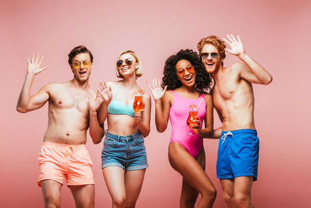 young multicultural friends in summer outfit waving hands at camera while holding cocktail glasses isolated on pink - Foto, imagen