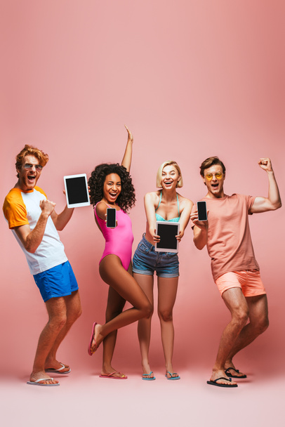 excited multicultural friends in summer outfit showing winner gesture while holding gadgets with blank screen on pink - Φωτογραφία, εικόνα