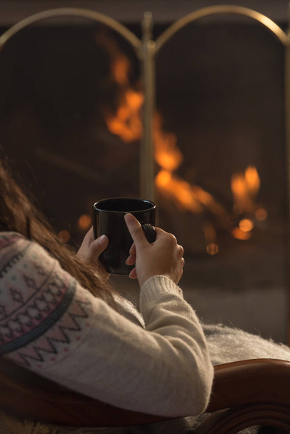 person with a wool sweater and long hair sitting in front of a lit fireplace and holding a black mug in his hands - Foto, Bild