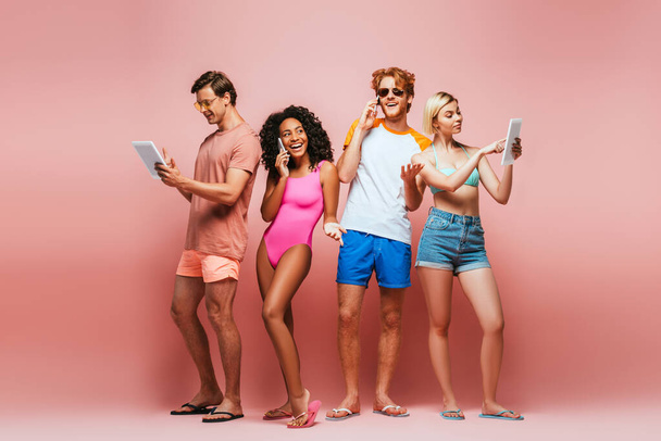 full length view of young multicultural friends in summer outfit using digital devices on pink - Foto, Imagen