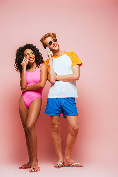 Multiethnic couple in sunglasses and swimsuit talking on smartphones on pink background - Fotografie, Obrázek