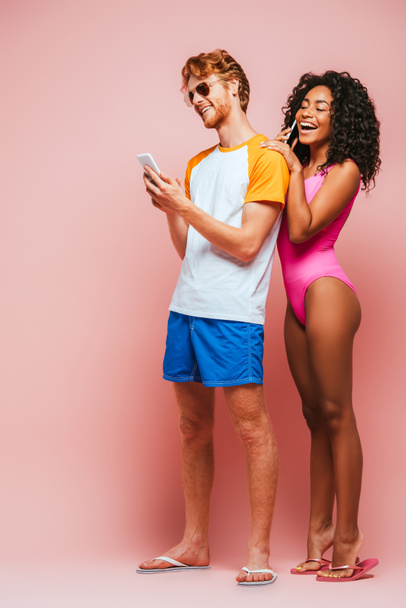 Femme afro-américaine en maillot de bain parlant sur smartphone près de l'homme en lunettes de soleil sur fond rose - Photo, image