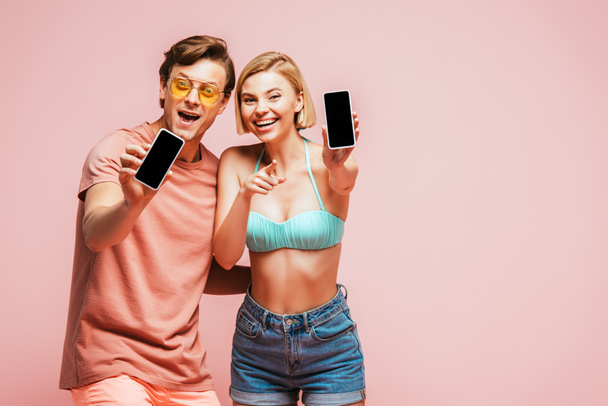 Young friends in sunglasses and swimsuit showing smartphones with blank screens and pointing with finger at camera isolated on pink  - Foto, Imagen