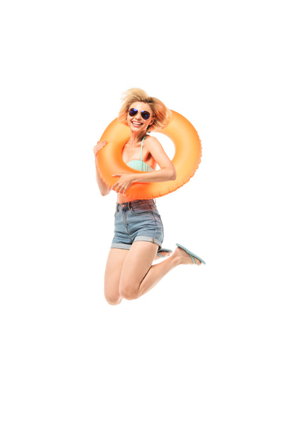 Mujer en gafas de sol y chanclas sosteniendo anillo de natación mientras salta aislado en blanco - Foto, Imagen