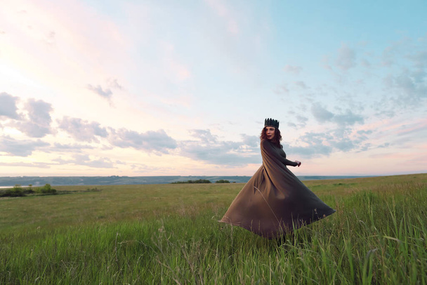 A young woman of 25-30 years old with curly red hair, in a green dress and a diadem with feathers, is spinning and dancing against the background of beautiful nature at dawn.  - Photo, Image