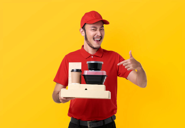 Азійський доставник, одягнений в червону форму, тримає коробку з-під обіду і каву, ізольовану на жовтому фоні. - Фото, зображення