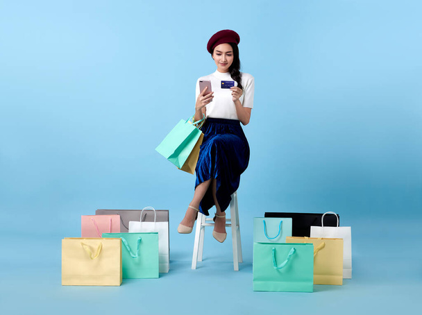 Азиатская покупательница сидит и несет сумки с кредитными картами и мобильным телефоном в руках на синем фоне. - Фото, изображение
