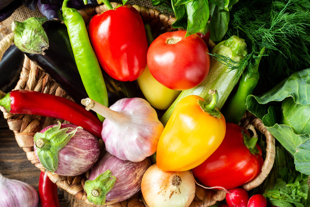 Various vegetables in a basket on a brown wooden table. Lots of raw vegetables in the basket. Eggplant, tomatoes, garlic, sweet peppers, onions on the table. Top view with space for text. Healthy food - Foto, imagen