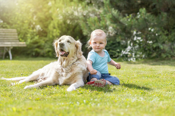 Cute liitle baby boy is posing with lying Golden Retriever dog on the grass - Foto, Imagen