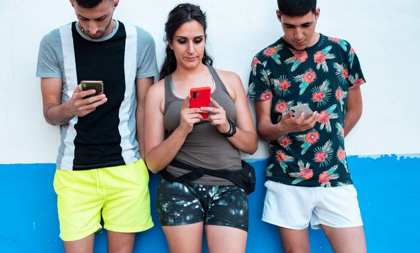 three young people, in summer clothes, on a white and blue wall, using their smartphones - Photo, Image