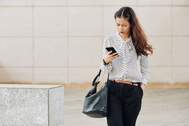 Serious joven mujer de negocios mixta caminando al aire libre y comprobando los mensajes de texto en su teléfono - Foto, Imagen