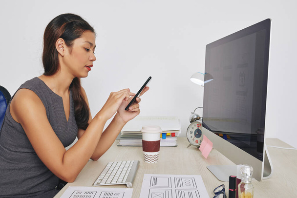 Junge UX-Designerin arbeitet am Schreibtisch an einem Projekt und liest Textnachrichten und E-Mails vom Kunden auf dem Smartphone-Bildschirm - Foto, Bild