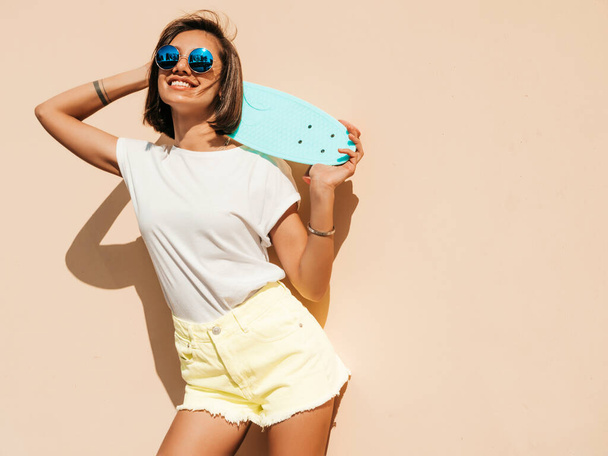 Young beautiful sexy smiling hipster woman in sunglasses.Trendy girl in summer T-shirt and shorts.Positive female with blue penny skateboard posing in the street near wall - Fotografie, Obrázek