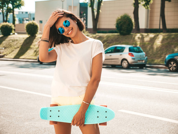 Young beautiful sexy smiling hipster woman in sunglasses.Trendy girl in summer T-shirt and shorts.Positive female with blue penny skateboard posing on the street background - Foto, imagen