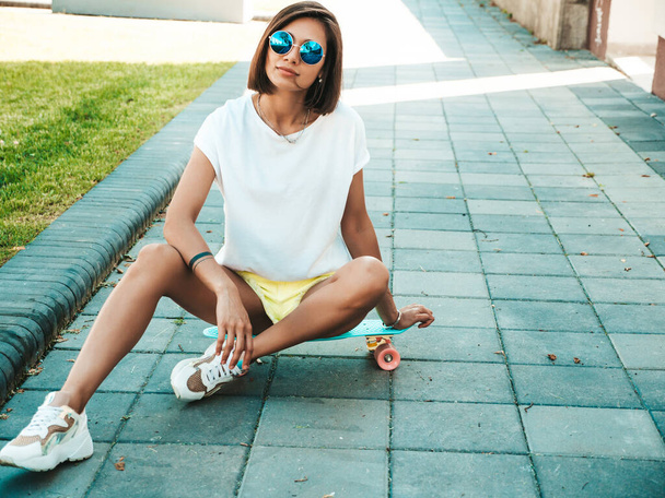 Young beautiful sexy smiling hipster woman in sunglasses.Trendy girl in summer T-shirt and shorts.Positive female with blue penny skateboard posing on the street background - Fotó, kép