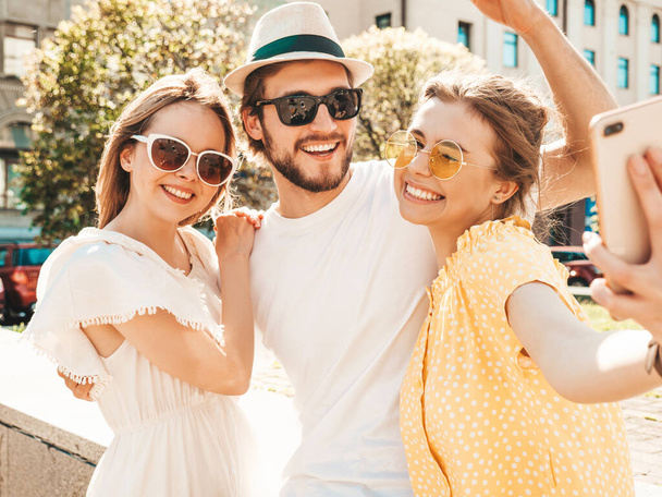 Group of young three stylish friends in the street.Man and two cute girls dressed in casual summer clothes.Smiling models having fun in sunglasses.Women and guy making photo selfie on smartphone - Foto, Imagen