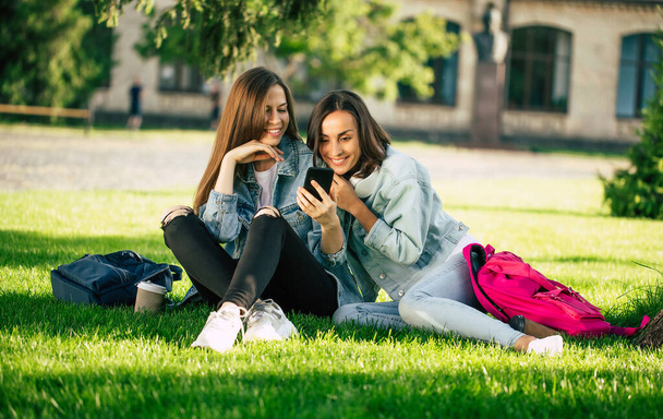 Zwei fröhliche und aufgeregte Studentinnen in lässiger Jeanskleidung entspannen mit Smartphones im College Park und haben Spaß. - Foto, Bild