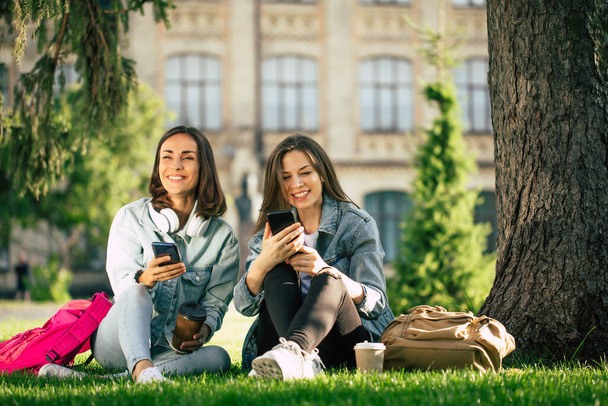 Two happy and excited student girl friends in casual denim clothes are relaxing in college park with smartphones and have fun. - Fotoğraf, Görsel