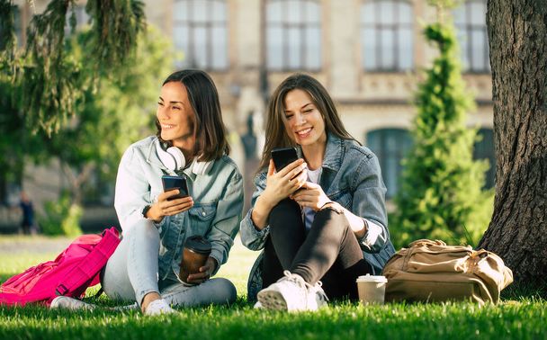 Two happy and excited student girl friends in casual denim clothes are relaxing in college park with smartphones and have fun. - Zdjęcie, obraz