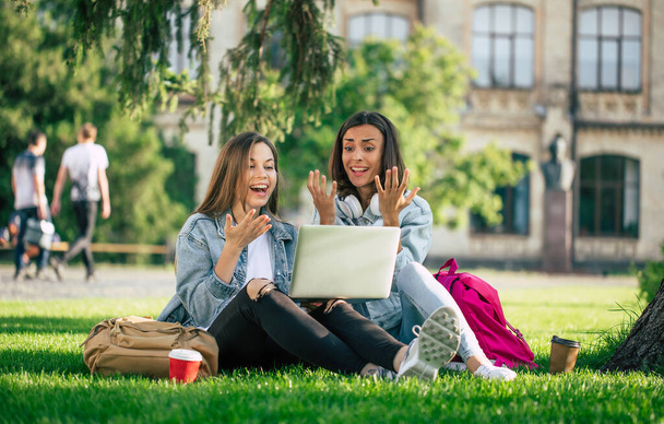 Two happy beautiful young student girl friends in casual denim clothes are relaxing in college park with laptop and smartphone on university background and drinking coffee. - Foto, Imagen