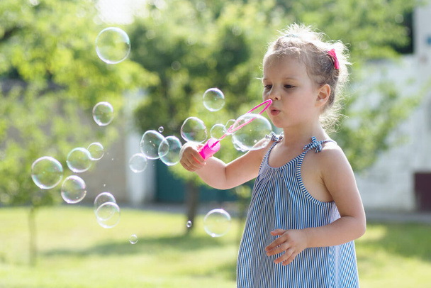 Cute little girl is blowing soap bubbles with pink bubble wand outdoors. - Фото, изображение