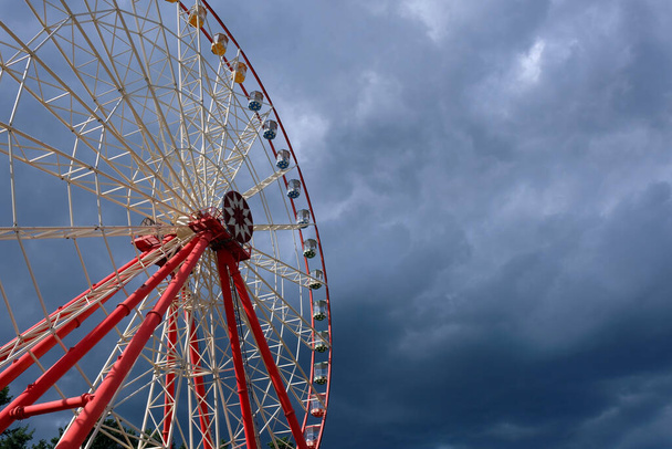 Красное колесо обозрения против серого облачного неба. Большое колесо обозрения в парке развлечений. Опасные достопримечательности. Закрытые кабины - Фото, изображение