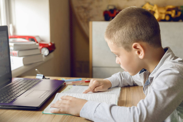 Хлопчик читає книжку, сидячи перед комп'ютером вдома. Робити домашнє завдання онлайн. Комфортне навчання
 - Фото, зображення