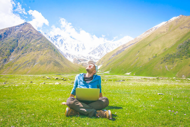 Мужчина среднего возраста сидит со скрещенными ногами на траве и наслаждается солнцем с серым ноутбуком на колене. Удаленная работа и самоизоляция в природе. - Фото, изображение