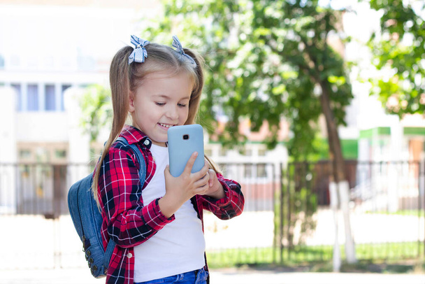 šťastná školačka s batohem, jak mluví po telefonu na ulici. v bílém tričku a kostkované košili - Fotografie, Obrázek