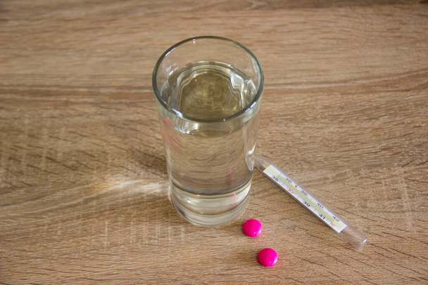 Un bicchiere d'acqua si trova sul tavolo mentre un termometro e pillole per il paziente si trovano accanto ad esso. Le compresse per il paziente si trovano sul tavolo con acqua. - Foto, immagini
