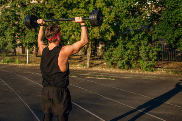 Male athlete lifting a barbell overhead, training athlete outdoors at sunset, sports equipment and clothing, free space for text, sun exposure, muscle building work, back view - Photo, Image