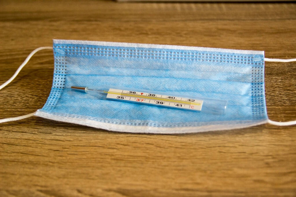 Thermometer voor het meten van de lichaamstemperatuur ligt in een masker ter bescherming tegen virussen. Een masker om te beschermen tegen het coronavirus ligt op tafel. - Foto, afbeelding