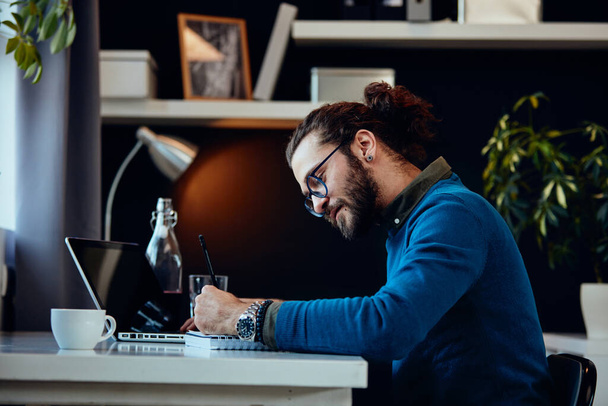 Side view of young attractive bearded freelancer sitting in his home office and writing tasks in notebook. - 写真・画像