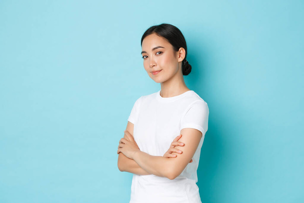 Portrait of happy and confident beautiful asian girl in white t-shirt cross arms chest and looking determined, standing half-turned over blue background, advertise product - Foto, Bild