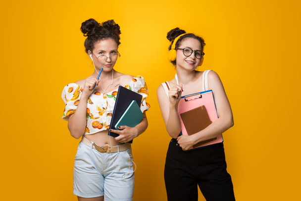 二人の白人女子大生が黄色のスタジオの壁にいくつかの本やフォルダを身に着けている眼鏡やイヤフォン - 写真・画像