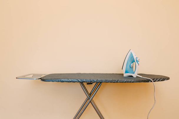 Uma tábua de engomar com um ferro azul-branco contra uma parede bege. - Foto, Imagem