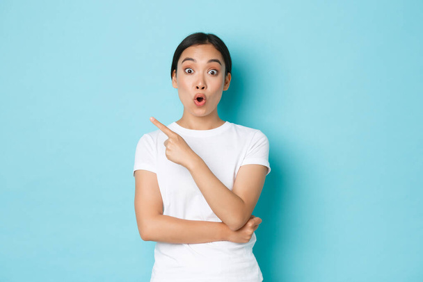 Πορτρέτο της έκθαμβης 20s όμορφο κορίτσι της Ασίας λαχανιάζοντας, κατσουφιάζοντας και λέγοντας wow, ενώ δείχνει πάνω αριστερή γωνία, δείχνοντας promo προσφορά, ζητώντας ερώτηση σχετικά με τη διαφήμιση, στέκεται μπλε φόντο - Φωτογραφία, εικόνα